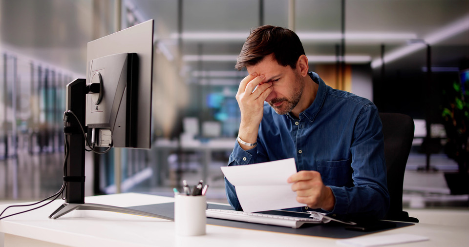 Stressed professional reading document at a modern office desk with computer setup.
