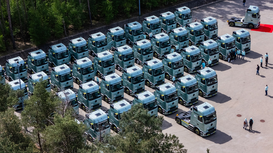 Flotte von grünen Lkw auf einem Parkplatz, umringt von Bäumen, bei einer Veranstaltung oder Präsentation.