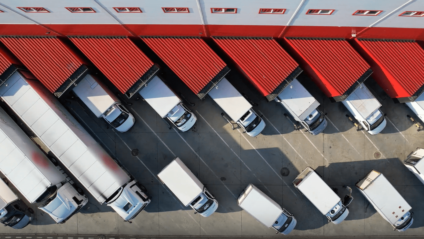 A yard with parked trucks on the right and a warehouse on the left, where several loading docks are open and trucks are being unloaded.