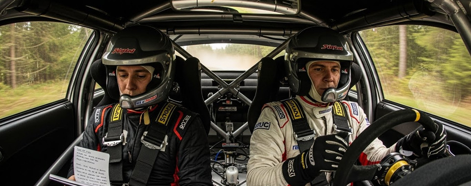 Two race car drivers in helmets, one reading a rally note, in a high-performance vehicle during a rally event.