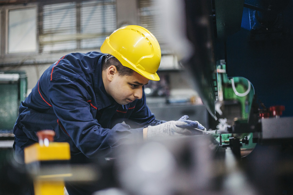 Ein Arbeiter in Schutzkleidung und Helm arbeitet an einer Maschine in einer Industrieumgebung.