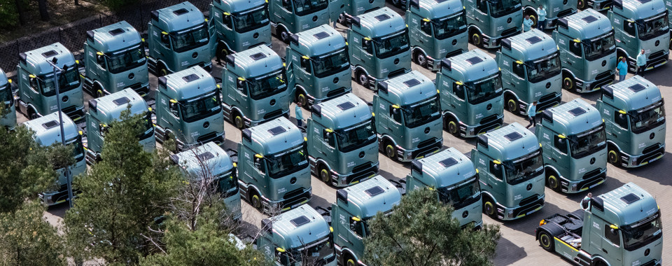 Aerial view of fleet of green electric trucks parked with people attending an event in a forested area.