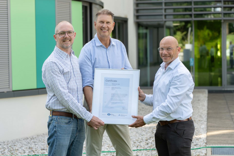 Three professionals proudly displaying a certification outside a modern building on a sunny day.