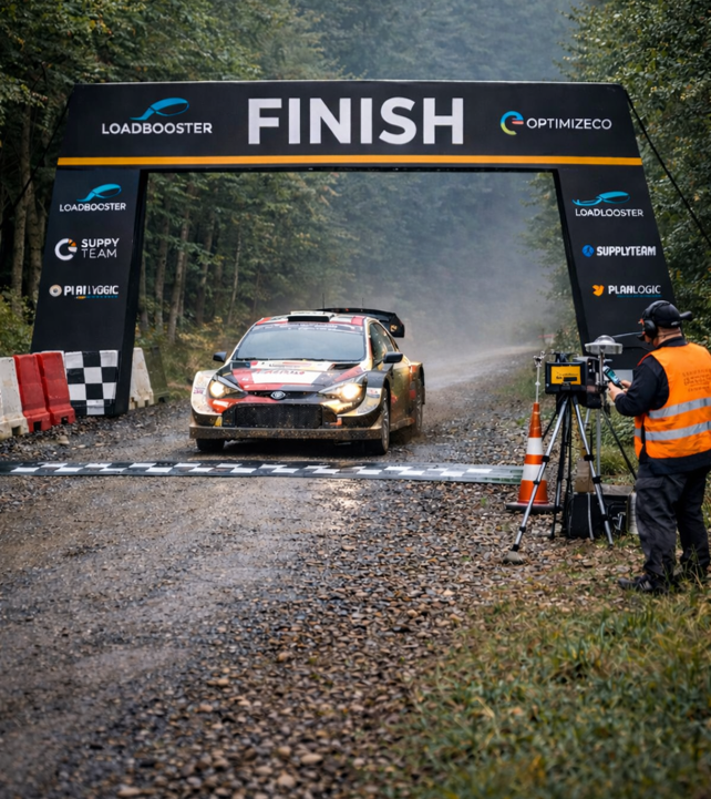 Rally car crossing the finish line at an event with spectators and timing equipment in a forest setting.