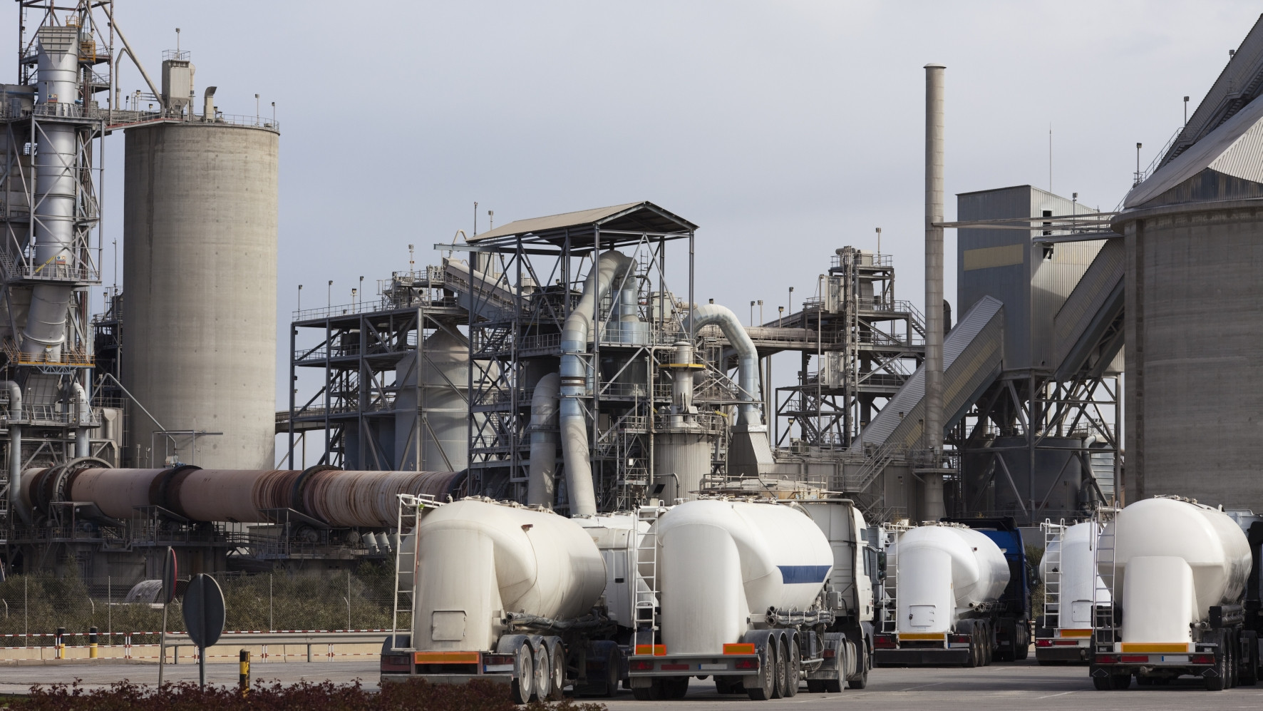 Industrial cement plant with large silos and concrete mixer trucks in the foreground on a cloudy day.