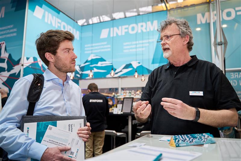 Menschen in einem Gespräch am INFORM-Stand auf einer Messe mit Unternehmensmaterialien.