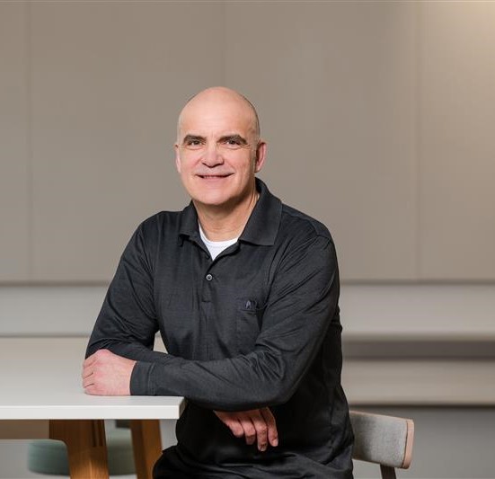 Portrait of a smiling man in a black polo shirt, seated at a table in a modern, bright workspace.