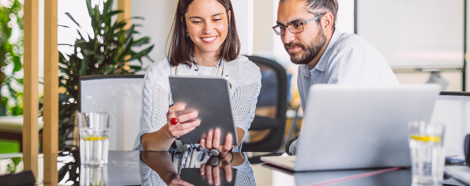 Two colleagues collaborating on a tablet in a modern office setting with plants and a laptop on the table.