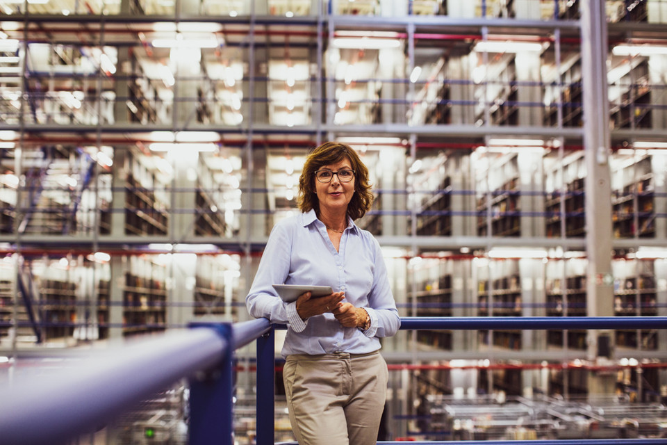 Professional woman in a warehouse holding a tablet, surrounded by tall storage shelves.