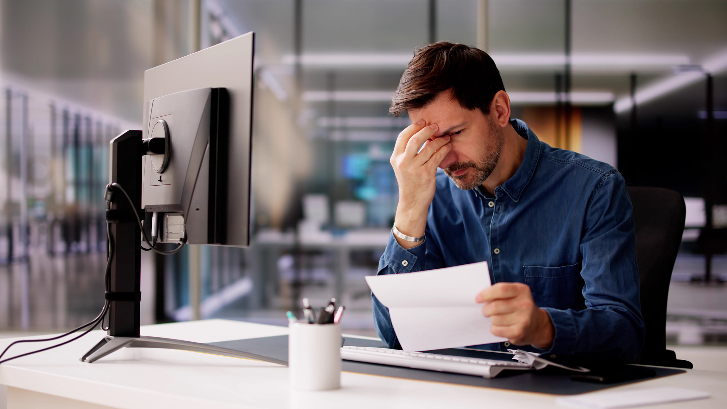 Stressed professional reading document at a modern office desk with computer setup.