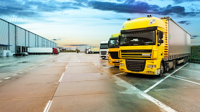 A yard with parked trucks on the right and a warehouse on the left, where several loading docks are open and trucks are being unloaded.