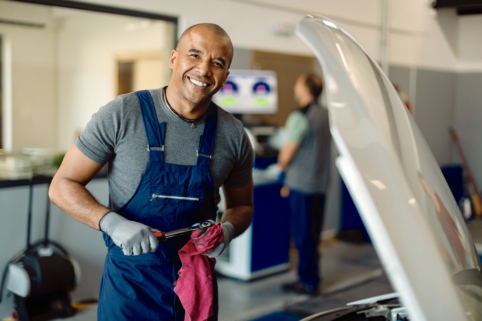 Automotive mechanic in blue overalls holding a wrench and red cloth, working on a vehicle in a workshop setting.