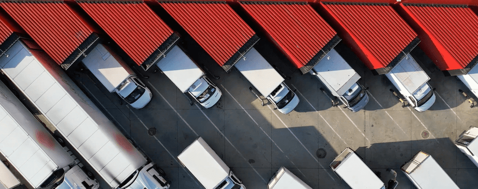 A yard with parked trucks on the right and a warehouse on the left, where several loading docks are open and trucks are being unloaded.