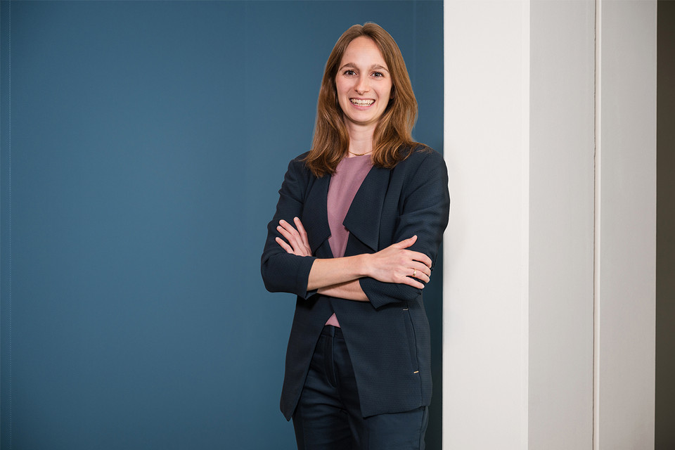 Smiling professional woman in a navy blazer, standing confidently against a blue wall, showcasing modern corporate space.