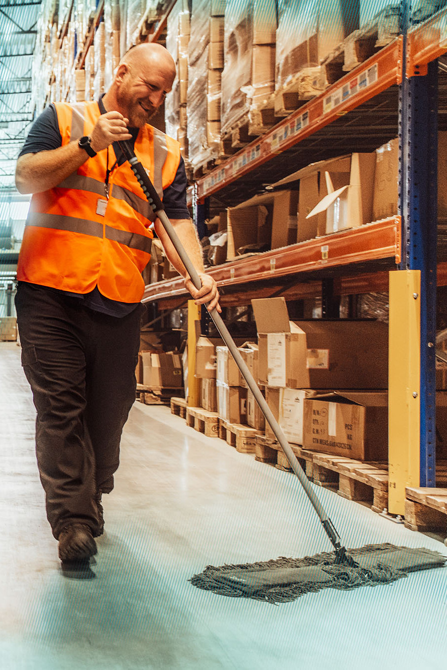 Warehouse employee cleaning aisle with mop, wearing safety vest, surrounded by stacked boxes and shelves.