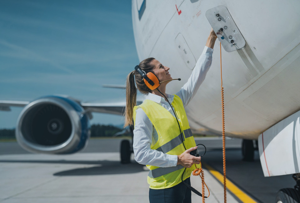 Female airline technician in safety vest using headset while inspecting an aircraft on the tarmac.