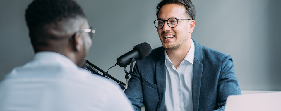 Zwei Männer bei einem Podcast-Interview mit Mikrofon und Laptop in einem modernen Raum.