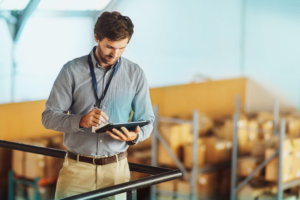 Warehouse manager using a tablet for inventory management among stacked cardboard boxes.