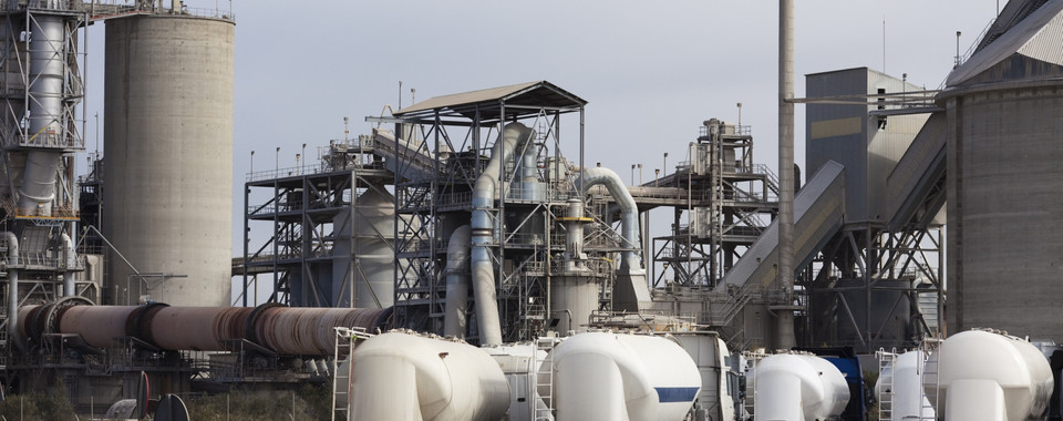 Industrial cement plant with large silos and concrete mixer trucks in the foreground on a cloudy day.
