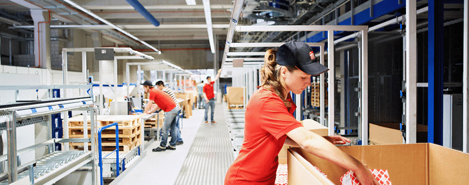 Workers in red shirts packaging items in a modern warehouse with organized pallets and efficient workspace design.
