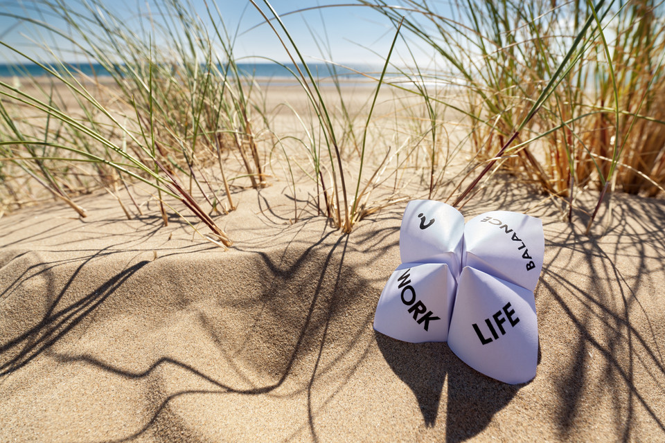 Papierobjekt im Sand zeigt die Begriffe Arbeit, Leben und Balance im Einklang mit der Natur am Strand.