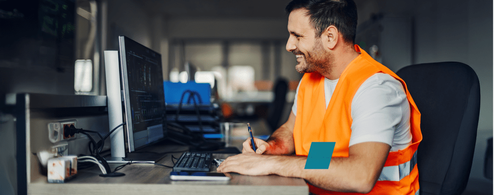 Smiling worker in an orange safety vest at a computer desk, focusing on digital projects in a modern office.