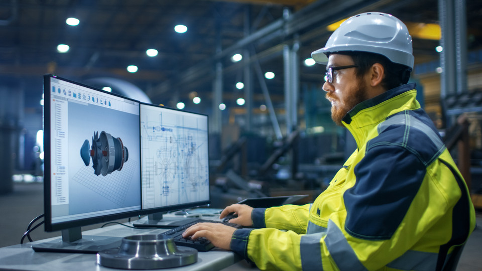 Ingenieur arbeitet am Computer mit 3D-Modellen in einer modernen Fabrikhalle mit technischer Ausstattung.