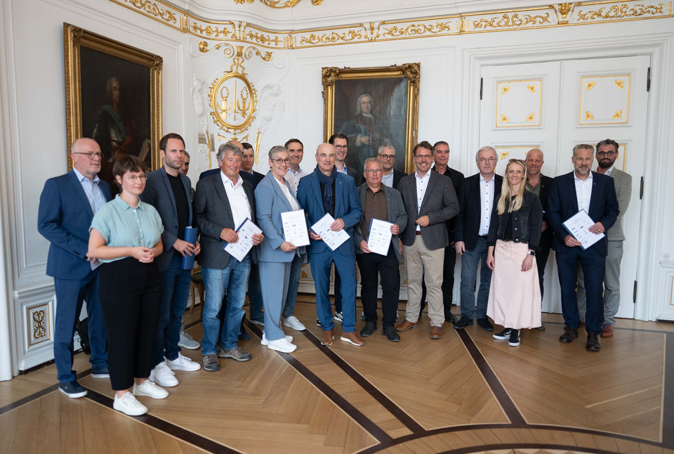 Gruppenfoto von Personen in formeller Kleidung mit Urkunden in elegantem Raum mit historischen Elementen.