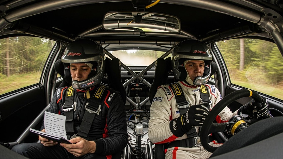 Interior view of a rally car with two drivers preparing for a race in helmets and racing gear.