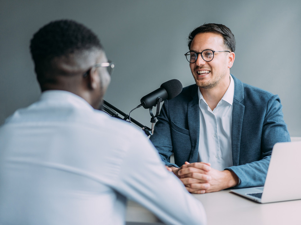 Zwei Männer bei einem Podcast-Interview mit Mikrofon und Laptop in einem modernen Raum.