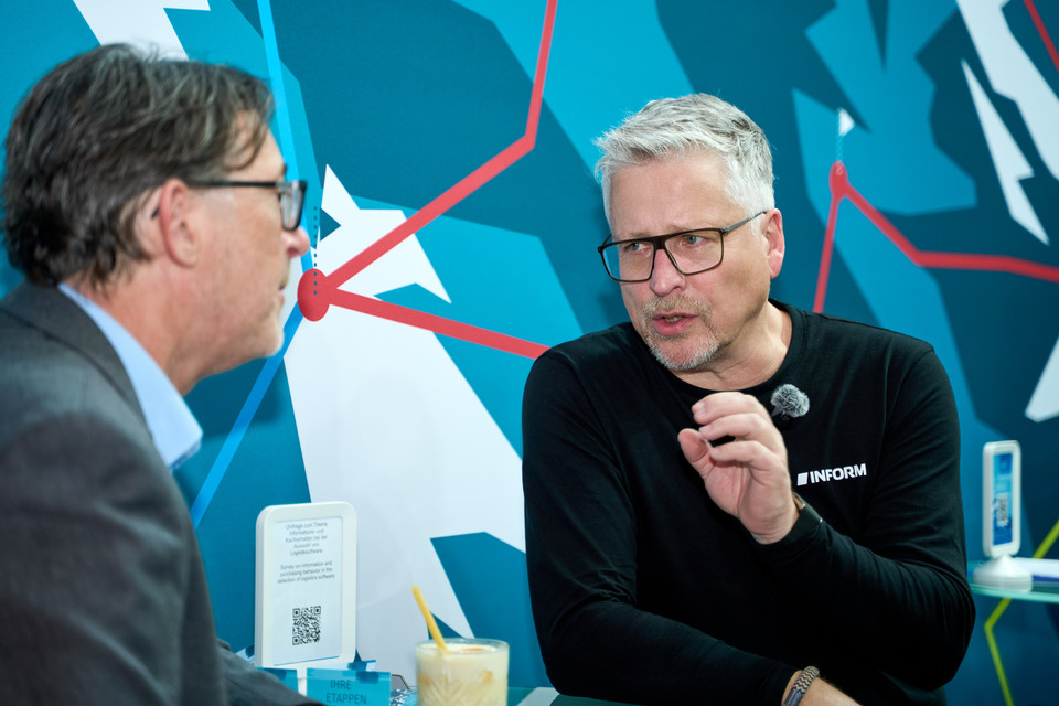 Zwei Männer in einem Gespräch am Stand mit abstraktem Hintergrund auf einer Messe. Fokus auf Interaktion und Austausch.