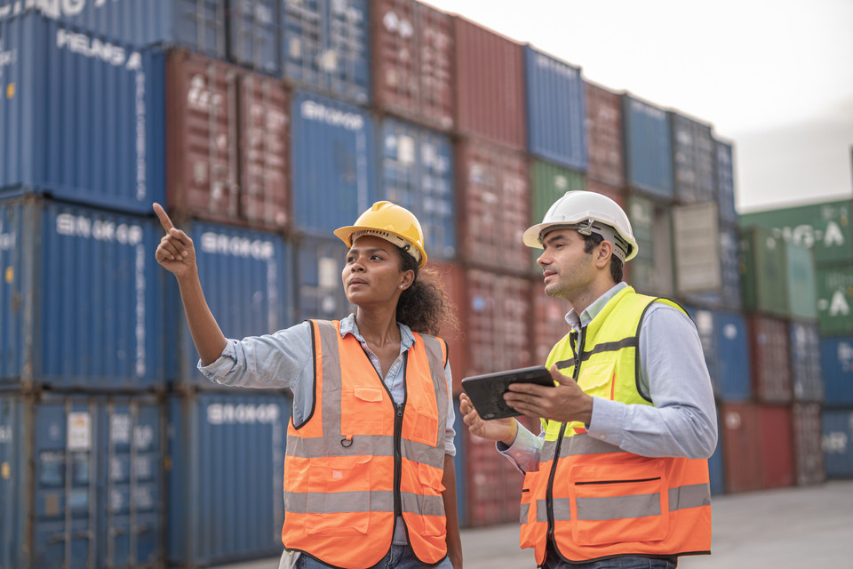 Two construction workers discussing logistics with shipping containers in the background at a port.