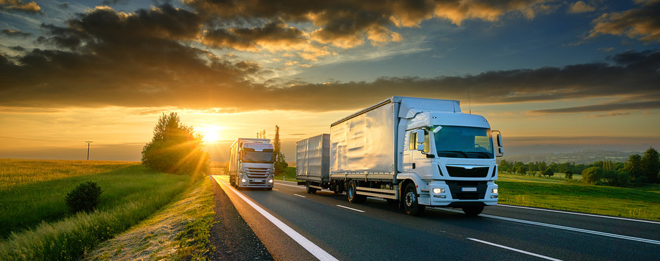 Two trucks driving on a deserted highway during a stunning sunset with golden skies and green fields.