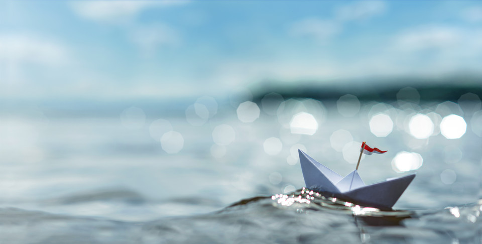 Ein kleines Papierboot mit roter Flagge schwimmt auf sparkling Wasser unter einem blauen Himmel.